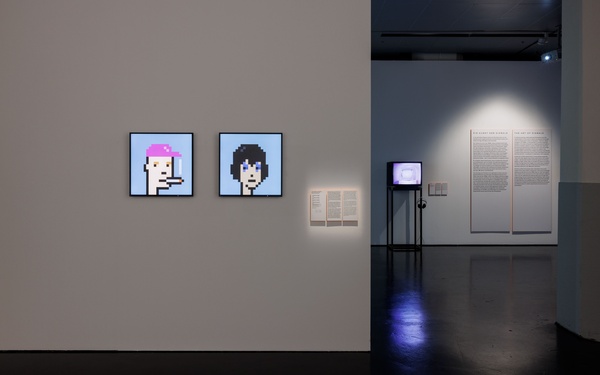 Blick in die Ausstellung mit zwei digitalen Bildern (NFTs) an einer weißen Wand, Infotafeln daneben und einem Monitor mit Kopfhörern im Hintergrund.