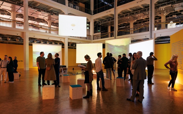 Visitors can be seen talking to each other in an exhibition space where video projections hang from the ceiling.