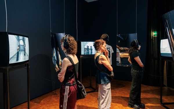 Mehrere Personen sind in einem Ausstellungsraum mit Bildschirmen und verzerrten Spiegeln zu sehen.