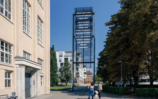Zu sehen sind zwei Personen und ein Hund, die sich eine Stehle mit Schrift vor blauem Himmel ansehen.