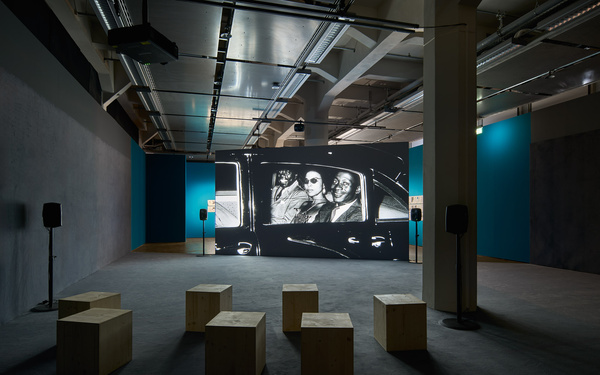 The picture shows an exhibition space with a large screen showing a black-and-white film. 