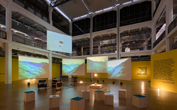The picture shows an exhibition space with several projection screens hanging from the ceiling. 