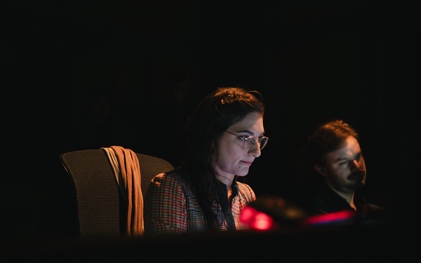 Zu sehen ist eine Person, die in einem dunkeln Raum sitzt und eine Brille trägt.  