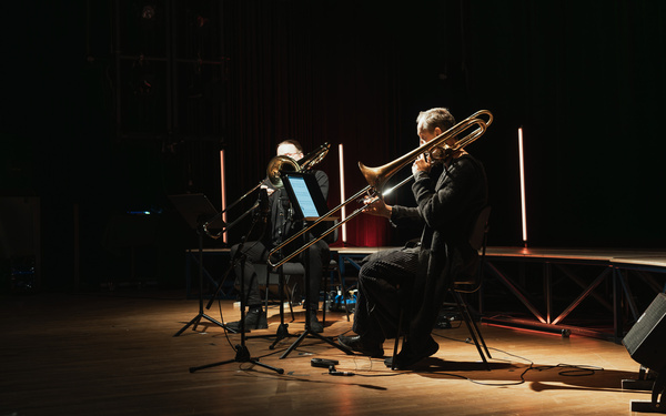 Zu sehen sind zwei Personen, die auf einer Bühne sitzen und Posaune spielen.