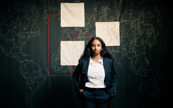 Amna Elamin steht vor einem schwarzen Stoffhintergrund mit Landkarten.