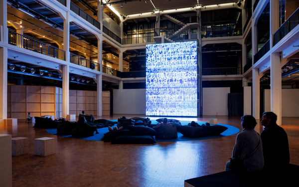 The scene shows a large screen with several people sitting on beanbag chairs in front of it. 
