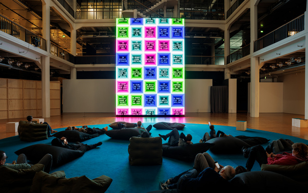 The scene shows a large screen with several people sitting on beanbag chairs in front of it. 