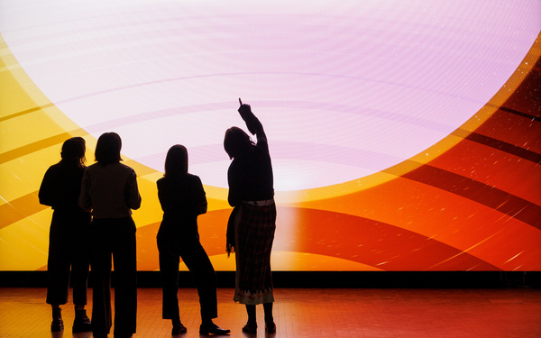 The image shows the silhouettes of four people standing in front of a giant screen. 