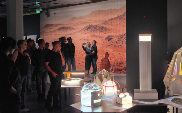 Zu sehen sind mehrere Personen in einem Ausstellungsraum, in dem Modelle auf Tischen präsentiert sind.