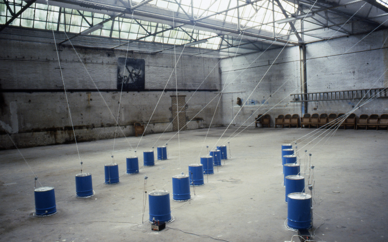 The picture shows an installation by Paul Panhuysen: Several dark blue barrels, from which ropes go to the ceiling, stand in a warehouse. 