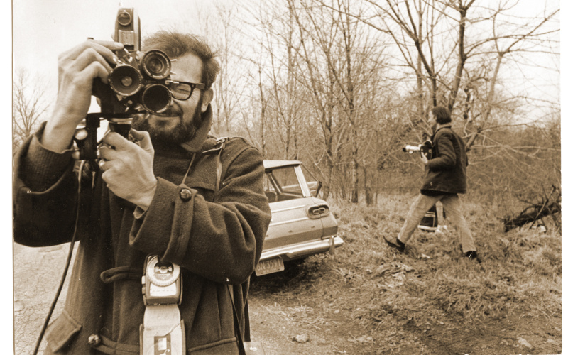 A black and white image of a man who also takes a photo. In the background a car and another man running in the picture