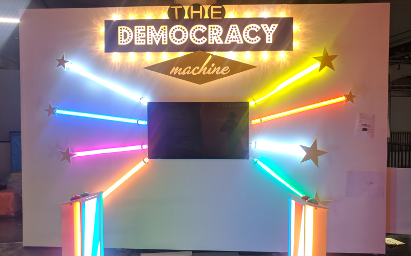 Front view of The Democracy Machine! In the middle of a wall is a television set installed around it, three colorful color tubes are arranged like sunbeams. In front of it are two podiums with buttons.