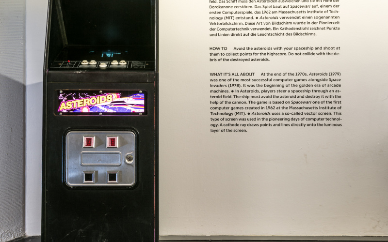 Ein »Asteroids« Arcade Automat vor einer Wand mit einem Beschreibungstext. 