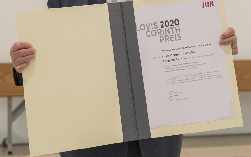 Ein Foto des Künstlers, Kurators und ZKM-Vorstands Peter Weibel, der den Lovis-Corinth Preis in Form einer Urkunde in den Händen hält.