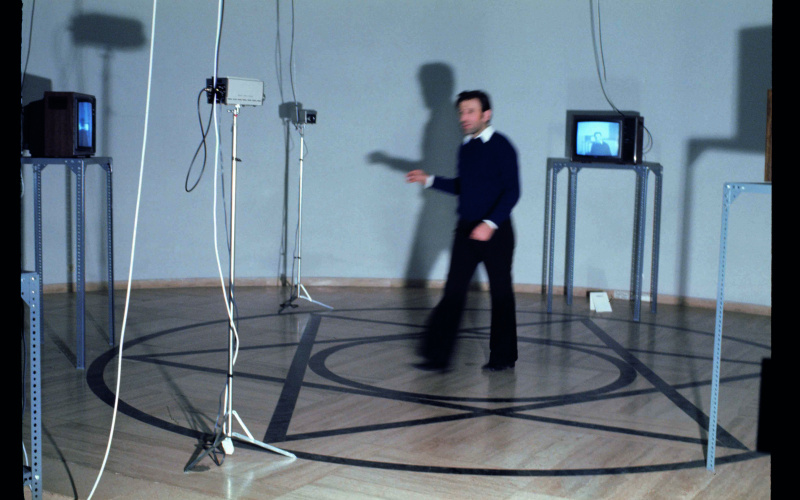 Ein Mann steht in einem Pentagramm umgeben von Monitoren und Kameras. Das Foto zeigt Peter Weibel im Rahmen der Ausstellung »respektive Peter Weibel«.