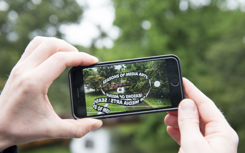 Zwei Hände halten ein Iphone auf dem ein App-Kunstwerk geöffnet ist. Nun sieht man die kleine Brücke im Zoo über der die Ausstellungs-Schlaufe schwebt.