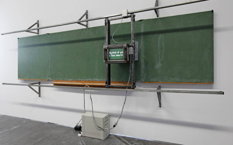 Ein Monitor auf Schienen vor einer grünen Tafel