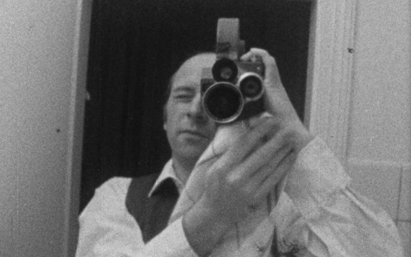 it is a black and white photo showing the artist Lutz Mommartz, with a large video camera over the shoulder, looking through it. 