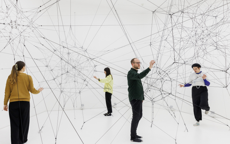 You can see a large net of threads in a white room. Several people are standing in this space and touching these threads.