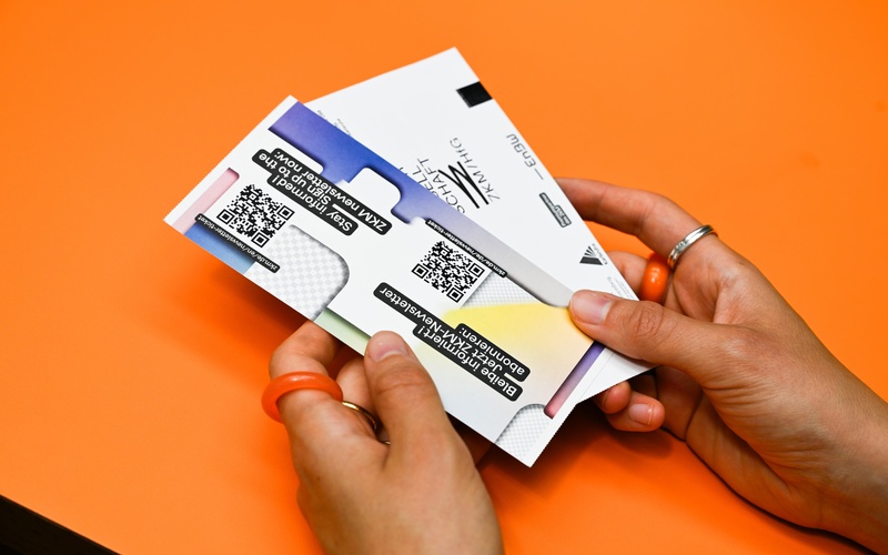 Zu sehen sind zwei Hände, die Eintrittskarten zum ZKM vor einen orangefarbenen Hintergrund halten. 
