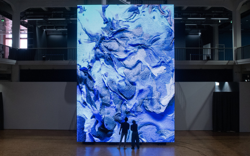 Zwei Silhouetten stehen vor einem riesigen Bildschirm, auf dem ein abstraktes Kunstwerk zu sehen ist.